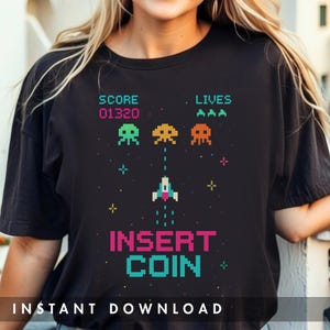 以下が含まれることがあります： レトロなビデオゲームのデザインが施された黒いTシャツ。デザインには、ピクセル化されたエイリアンのような生き物、宇宙船、「SCORE 01320」、「LIVES AAA」、「INSERT COIN」の文字が含まれています。このシャツはヴィンテージ感があります。
