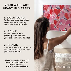 May include: A woman hangs a framed print of a pink and red heart design on a white wall. The print is titled "Fern Prints". The text "Your Wall Art Ready in 3 Steps" is on the left side of the image. The steps are "Download", "Print", and "Frame".