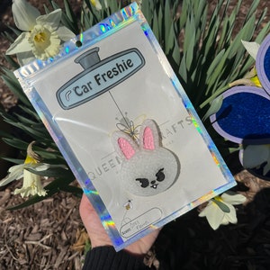 May include: A white bunny car freshener with pink ears and black eyes. The bunny is in a clear plastic bag with a white label that says "Car Freshie" and "Queen Bee Crafts". The label also says "Scent: Baby Powder".