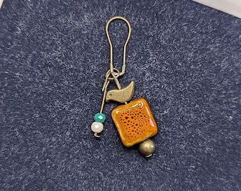 May include: A decorative stitch marker with a gold-colored hook and a square, orange-brown bead with a speckled pattern. A small bird charm and a cluster of white, teal, and gray beads add detail. The marker is designed for knitting or crochet projects.