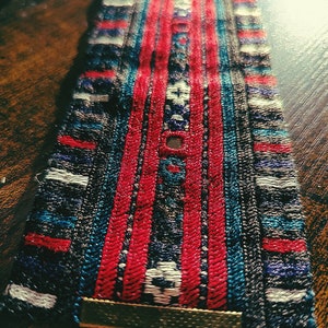 May include: A close-up of a colorful woven bracelet with red, blue, and white stripes. The bracelet has a metal clasp and a brown leather strap.