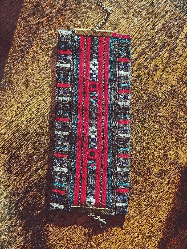 May include: A woven fabric bracelet with a chain and clasp closure. The bracelet features a colorful geometric pattern in shades of red, blue, and gray.