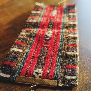 May include: A colorful woven bracelet with a gold metal clasp and a silver hook closure. The bracelet has a geometric pattern with red, blue, and white stripes.