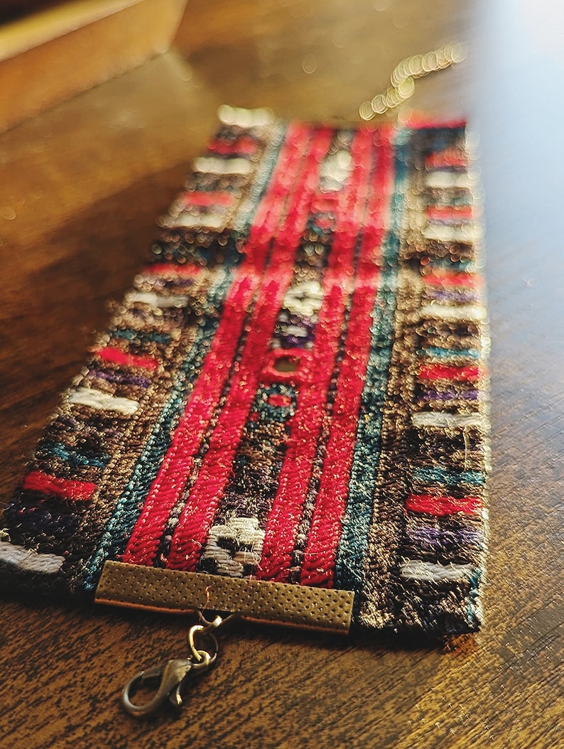 May include: A colorful woven bracelet with a gold metal clasp and a silver hook closure. The bracelet has a geometric pattern with red, blue, and white stripes.