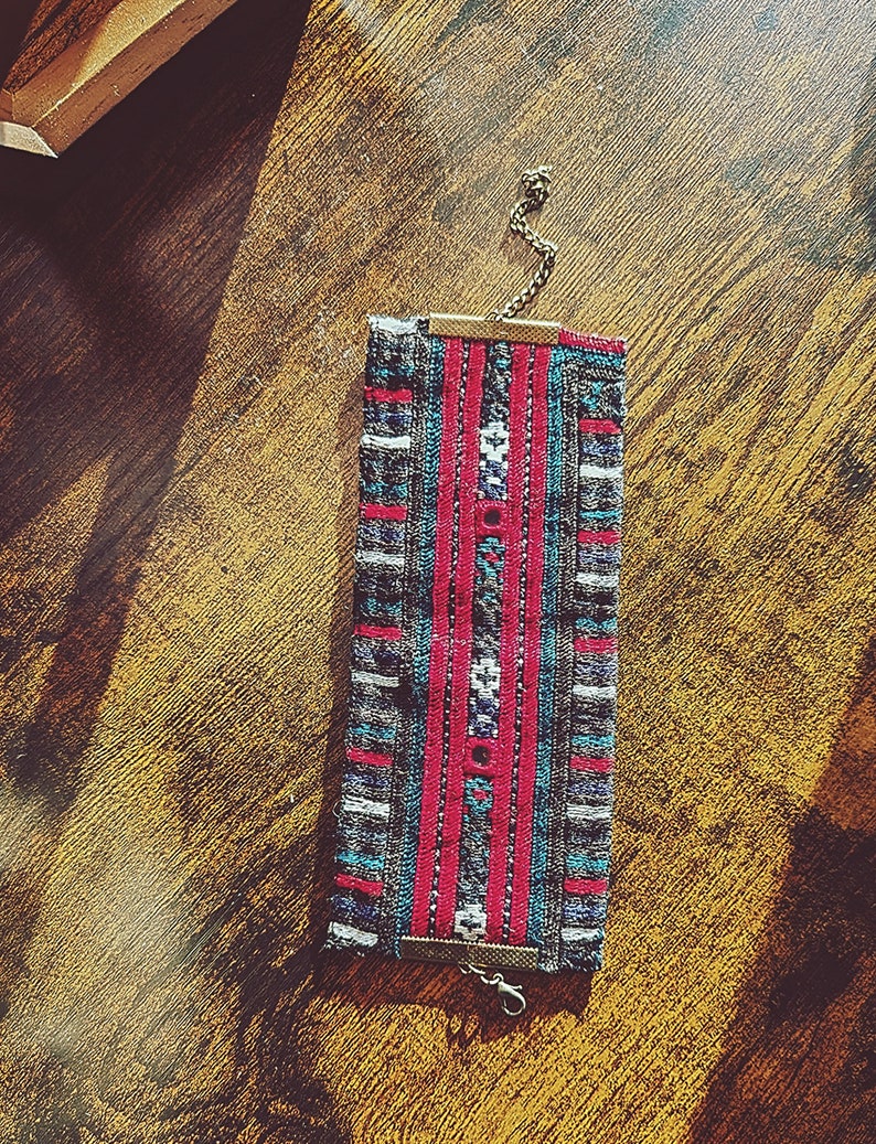 May include: A woven fabric bracelet with a geometric pattern in shades of gray, blue, red, and white. The bracelet has a metal clasp and chain.