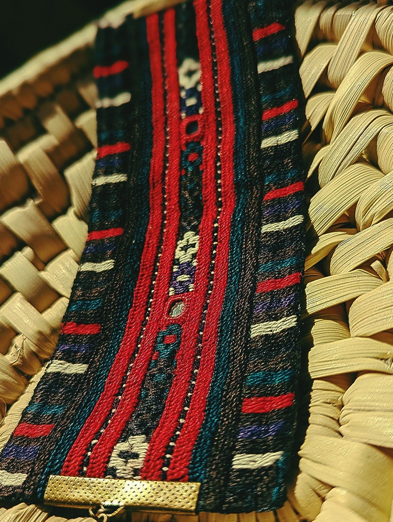 May include: A woven bracelet with a red, blue, and white geometric pattern. The bracelet has a gold metal clasp.
