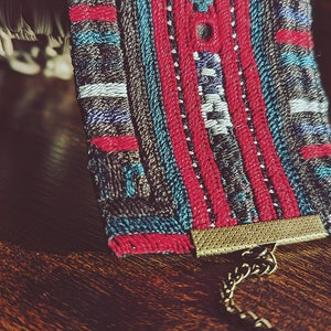 May include: A colorful woven fabric bracelet with a gold chain and clasp. The bracelet features a geometric pattern in shades of red, blue, and white.