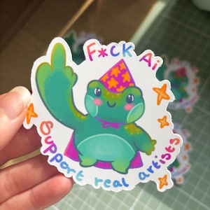 May include: A sticker with a cartoon frog giving the middle finger. The frog is wearing a party hat and cape, with text that reads "F*ck Ai Support real artists" in a rainbow font. Orange star accents surround the frog.