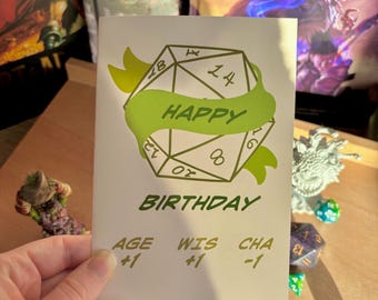 Puede incluir: Una tarjeta de cumpleaños verde y blanca con un dado verde de 20 caras y el texto "Feliz cumpleaños". La tarjeta también tiene el texto "Age +1, Wis +1, Cha -1".