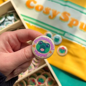 May include: A circular badge featuring a cartoon frog and the text "Professional Yapper". The badge is held up, with other badges and a yellow background with orange text visible. The background has the words "Cosy Spu".