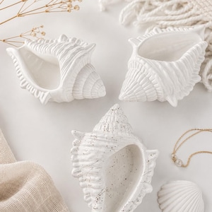 Peut inclure: Trois bols en céramique en forme de coquillage blanc. Deux bols ont une finition blanche unie et lisse avec des crêtes détaillées, tandis que le troisième présente une surface blanche avec de petites taches noires. Chaque coquille est creuse.