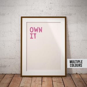 Op de afbeelding: Ingelijste kunstdruk met de woorden "OWN IT" in roze op een lichtroze achtergrond. De print zit in een bruine lijst en leunt tegen een witte muur en een houten vloer.