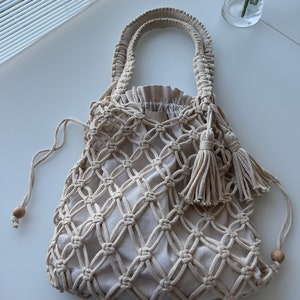 Peut inclure: Un sac cabas en crochet beige avec une fermeture à cordon et des accents de perles en bois. Le sac a un design noué et deux longues poignées.