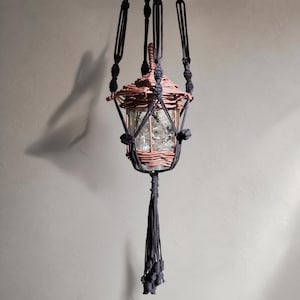 May include: A hanging lantern featuring a clear glass jar encased in a brown woven wicker frame. The lantern is suspended by black macrame cord with knotted details and a long, decorative tassel hanging from the bottom center of the structure.