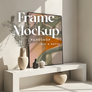 Könnte beinhalten: Ein weißes Regal mit einem schwarzen gerahmten Druck von abstrakter Kunst mit grünen und rosa Farben. Der Druck ist mit "Frame Mockup Photoshop DIN A Ratio" betitelt. Auf dem Regal stehen drei kleine dekorative Vasen und eine größere Vase mit einer Pflanze auf der linken Seite des Regals.