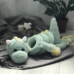 May include: A light green crocheted dragon toy with yellow detailing on its feet and head. The dragon features white horns and a long, coiled body. The toy is resting on a grey surface.