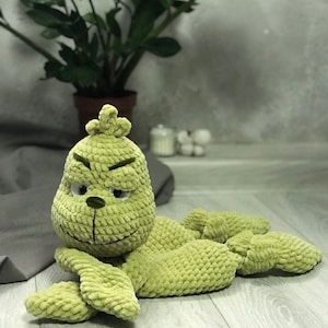 Puede incluir: Muñeco Grinch tejido a ganchillo en verde claro con expresión gruñona. El peluche tiene una superficie texturizada y está tumbado sobre una superficie de madera. Los brazos y las piernas del Grinch están extendidos, y el muñeco está hecho de hilo suave.