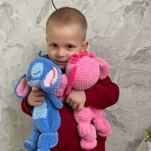 May include: Two plush toys: a blue creature with large ears and a pink creature with a bow. The blue toy has pink inner ears and a stitched mouth. The pink toy has a pink bow. Both toys are held in front of a person wearing a red jumper.
