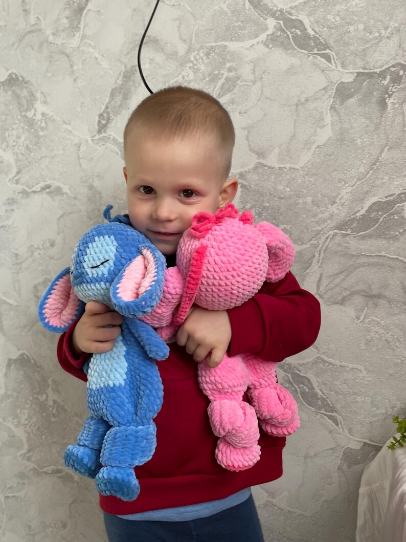 May include: Two plush toys: a blue creature with large ears and a pink creature with a bow. The blue toy has pink inner ears and a stitched mouth. The pink toy has a pink bow. Both toys are held in front of a person wearing a red jumper.