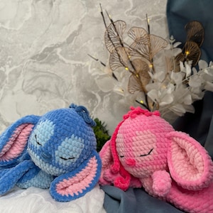 May include: Two crocheted bunny toys in blue and pink. The blue bunny has pink accents, and both have closed eyes and long ears. They are lying down. The background features a marble-effect wall and decorative floral arrangements.