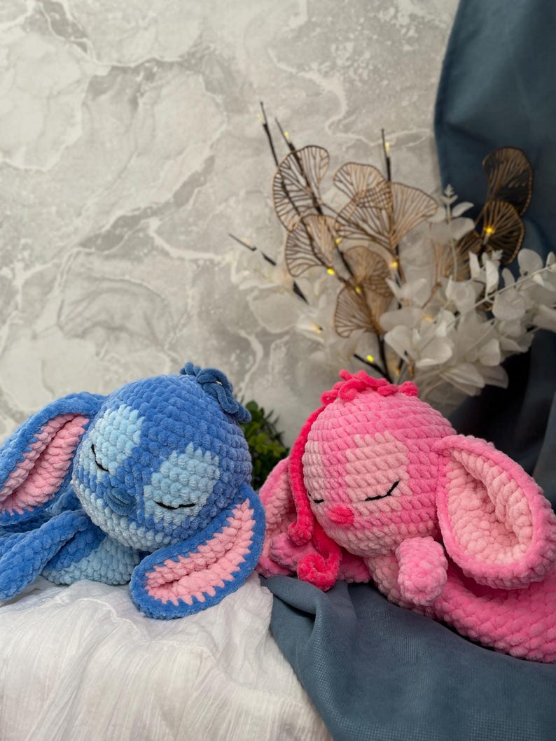 May include: Two crocheted bunny toys in blue and pink. The blue bunny has pink accents, and both have closed eyes and long ears. They are lying down. The background features a marble-effect wall and decorative floral arrangements.