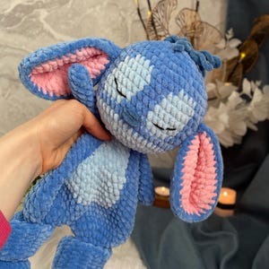 May include: A handmade blue plush toy with pink inner ears and a light blue belly patch. The toy has closed eyes and a textured, crocheted appearance, resembling a cartoon character. The plush is held in a person's hand.