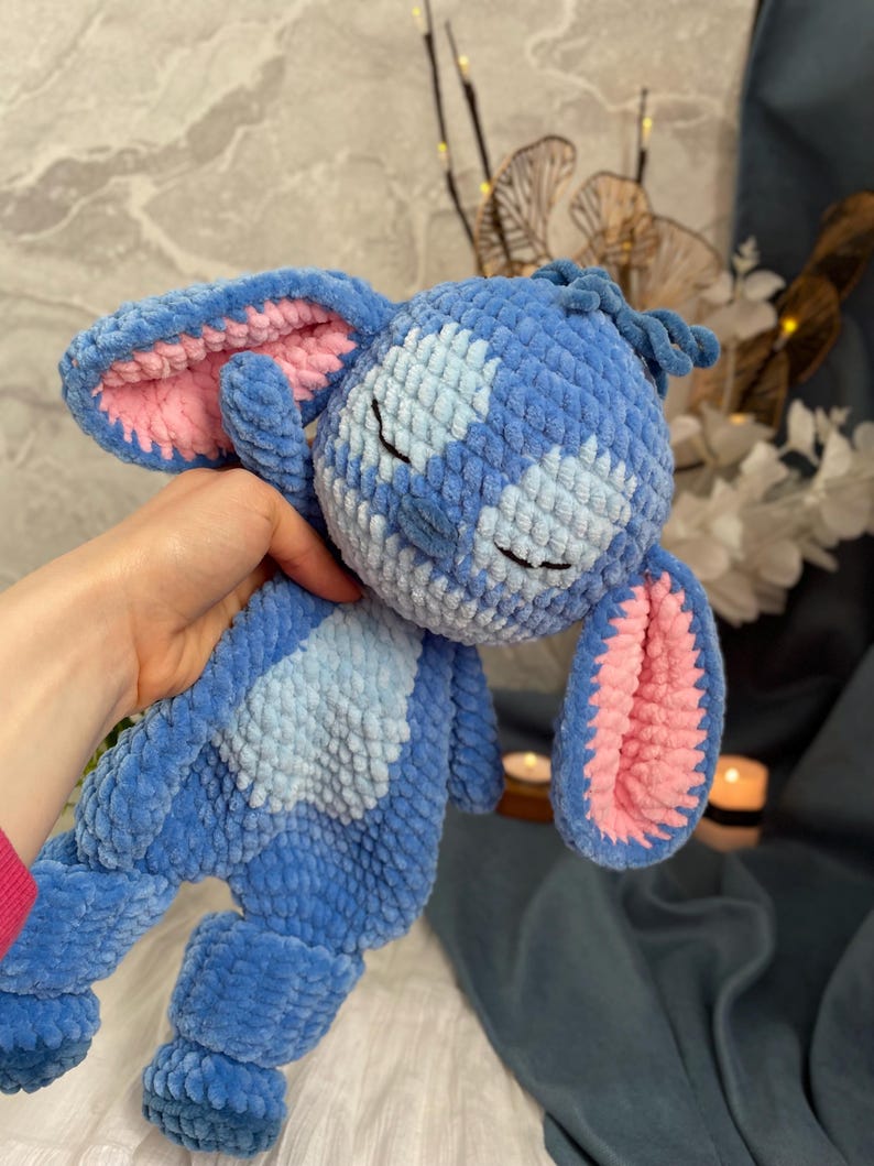 May include: A handmade blue plush toy with pink inner ears and a light blue belly patch. The toy has closed eyes and a textured, crocheted appearance, resembling a cartoon character. The plush is held in a person's hand.
