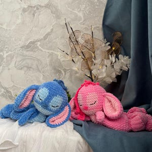 May include: Two handmade crochet bunny plushies. One is blue with pink inner ears, the other is pink with pink inner ears. Both are lying down with closed eyes, suggesting sleep. The bunnies are on a white surface with a blue backdrop.