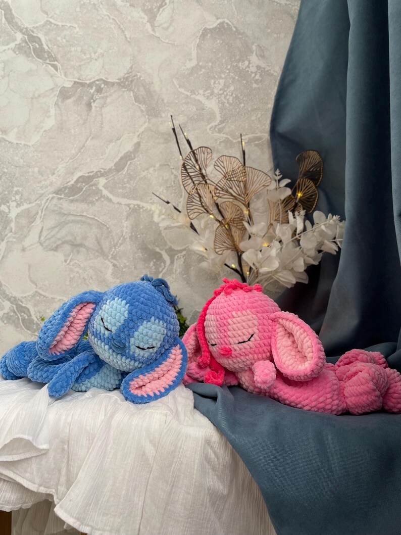 May include: Two handmade crochet bunny plushies. One is blue with pink inner ears, the other is pink with pink inner ears. Both are lying down with closed eyes, suggesting sleep. The bunnies are on a white surface with a blue backdrop.