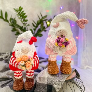 May include: Two crocheted gnomes, one in red and white and the other in pink and white, holding bouquets of flowers. The gnomes are standing on a white and black plaid blanket.