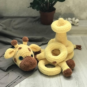 Peut inclure: Un jouet en crochet en forme de girafe jaune avec des taches brunes et un nez brun. La girafe tient un jouet en anneau en crochet jaune. Il y a un jouet d'empilage en crochet jaune derrière la girafe.