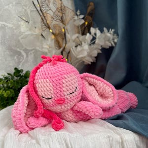 May include: A handmade pink crochet bunny plush toy. The bunny is lying down with its eyes closed, featuring long floppy ears and a small pink nose. The plush is made of soft, textured yarn.