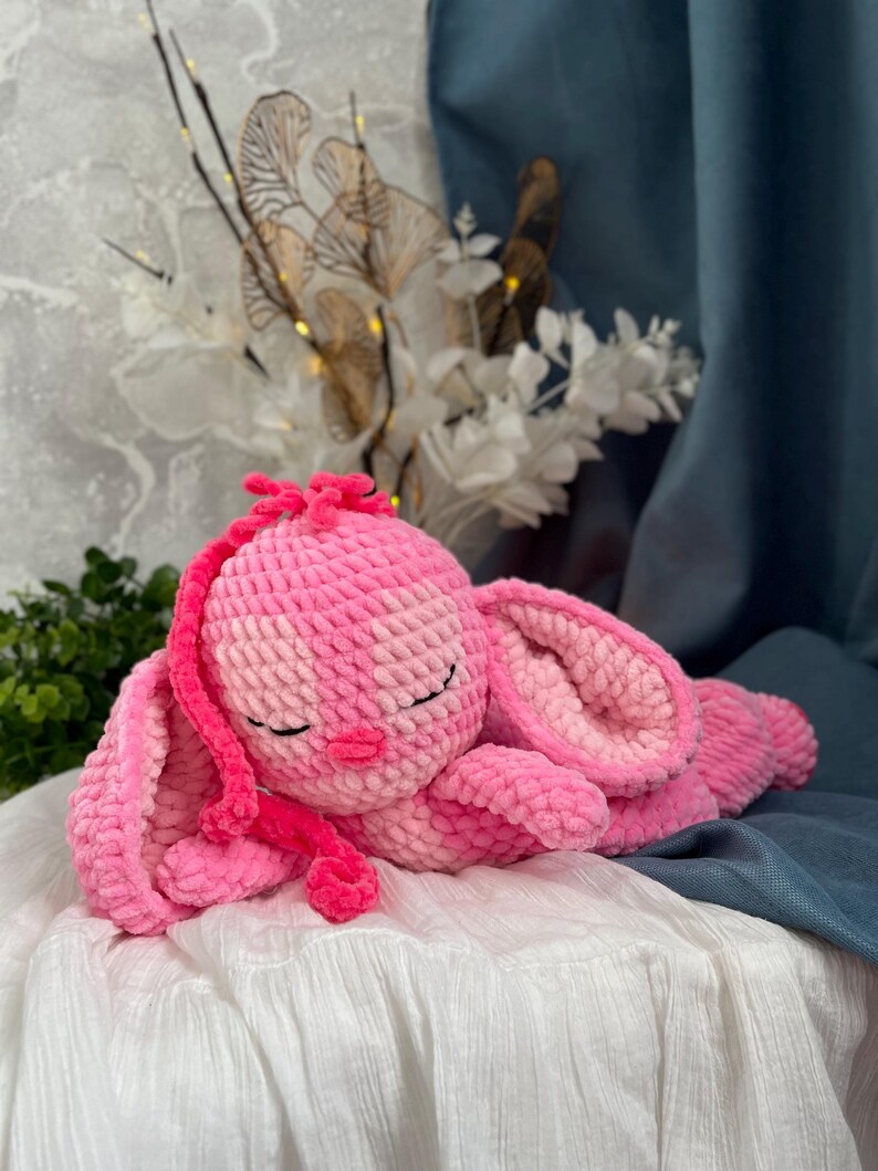 May include: A handmade pink crochet bunny plush toy. The bunny is lying down with its eyes closed, featuring long floppy ears and a small pink nose. The plush is made of soft, textured yarn.