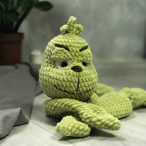 May include: Green crocheted plush toy resembling the Grinch from the movie How the Grinch Stole Christmas. The toy has a grumpy expression with large eyes and a wide mouth.