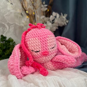 May include: A handmade pink crochet bunny plush toy with closed eyes, long ears, and a small pink nose. The plush is lying on a white surface, with a soft, textured appearance. The bunny is a light and dark pink colour.