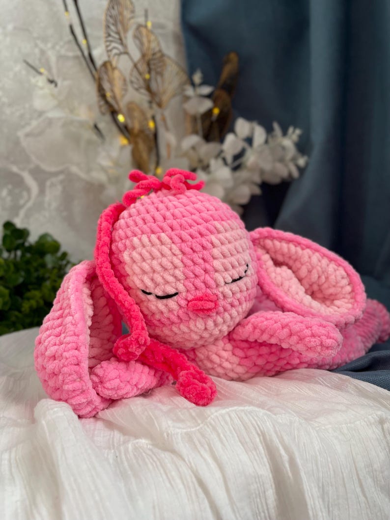 May include: A handmade pink crochet bunny plush toy with closed eyes, long ears, and a small pink nose. The plush is lying on a white surface, with a soft, textured appearance. The bunny is a light and dark pink colour.