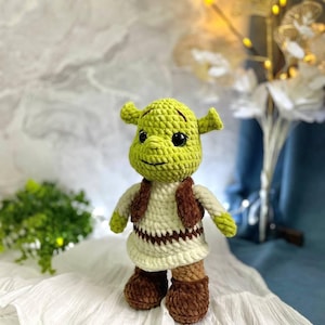Op de afbeelding: Een groen gehaakt pluche speelgoed van Shrek, een personage uit de Shrek-filmfranchise. Het speelgoed draagt een bruin vest en een witte jurk met bruine bies. Het speelgoed staat op een wit gehaakt oppervlak.