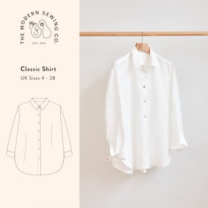 以下が含まれることがあります： ボタンダウンカラーと長袖の白いリネンシャツ。シャツは木製ハンガーにかけられています。画像の下には「Classic Shirt」と「UK Sizes 4 - 28」のテキストが印刷されています。