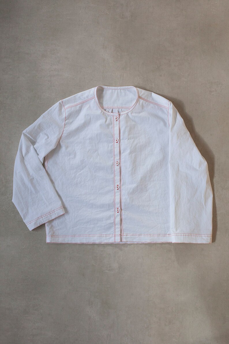 Pu&ograve; includere: Una camicia bianca a maniche lunghe con chiusura a bottoni sul davanti e cuciture rosse sui bordi. La camicia ha un collo rotondo e una vestibilit&agrave; rilassata.
