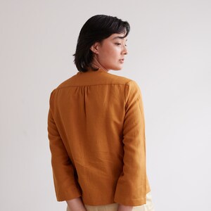 Pu&ograve; includere: Una donna indossa una camicia di lino color senape con maniche a tre quarti. La camicia ha una vestibilit&agrave; rilassata e una lunghezza leggermente corta. La donna indossa pantaloni beige e ha le mani giunte dietro la schiena.