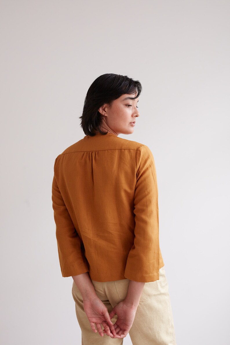 Pu&ograve; includere: Una donna indossa una camicia di lino color senape con maniche a tre quarti. La camicia ha una vestibilit&agrave; rilassata e una lunghezza leggermente corta. La donna indossa pantaloni beige e ha le mani giunte dietro la schiena.
