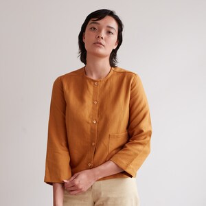 Pu&ograve; includere: Una persona indossa una camicia di lino color senape con chiusura a bottoni sul davanti e una sola tasca sul lato sinistro. La camicia ha maniche lunghe che sono arrotolate fino ai gomiti.