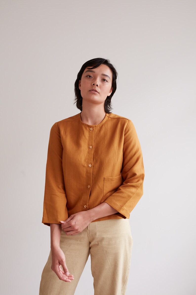 Pu&ograve; includere: Una persona indossa una camicia di lino color senape con chiusura a bottoni sul davanti e una sola tasca sul lato sinistro. La camicia ha maniche lunghe che sono arrotolate fino ai gomiti.