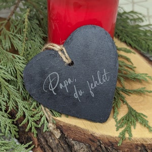 May include: A black heart-shaped slate plaque with the text "Papa, du fehlst" engraved on it. The plaque is sitting on a wooden surface with a red candle holder in the background.