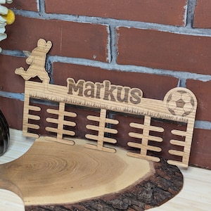 Könnte beinhalten: Medaillenhalter aus Holz mit einem Fußballspieler und einem Fußball. Der Halter hat 12 Fächer für Medaillen und der Name "Markus" ist oben eingraviert.
