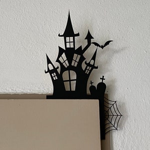Może przedstawiać: Czarna dekoracja narożna na Halloween z sylwetką nawiedzonego domu z nietoperzem, nagrobkami i pajęczyną. Dekoracja wykonana jest z ciemnego materiału i jest przeznaczona do umieszczenia w rogu.