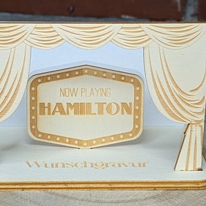 Peut inclure: Un accessoire de sc&egrave;ne en bois avec une enseigne de cin&eacute;ma qui indique "NOW PLAYING HAMILTON". La sc&egrave;ne est d&eacute;cor&eacute;e de rideaux et les mots "Wunschgravur" sont imprim&eacute;s sur la base.