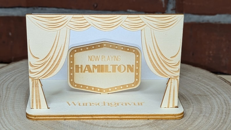 Peut inclure: Un accessoire de sc&egrave;ne en bois avec une enseigne de cin&eacute;ma qui indique "NOW PLAYING HAMILTON". La sc&egrave;ne est d&eacute;cor&eacute;e de rideaux et les mots "Wunschgravur" sont imprim&eacute;s sur la base.