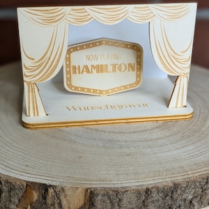 Peut inclure: Une sc&egrave;ne en bois avec une enseigne de cin&eacute;ma qui indique "NOW PLAYING HAMILTON". La sc&egrave;ne est d&eacute;cor&eacute;e de rideaux et les mots "Wunschgravur" sont imprim&eacute;s sur la base.