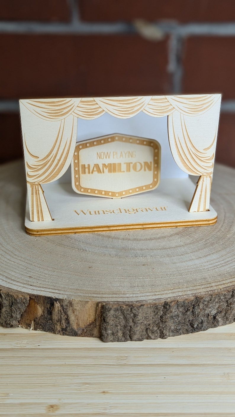 Peut inclure: Une sc&egrave;ne en bois avec une enseigne de cin&eacute;ma qui indique "NOW PLAYING HAMILTON". La sc&egrave;ne est d&eacute;cor&eacute;e de rideaux et les mots "Wunschgravur" sont imprim&eacute;s sur la base.